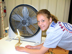 A girl stands in front of a fan with a gold wind turbine in front of it.