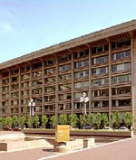 Photo of the exterior front of 950 L'Enfant Plaza. The entryway is lined with a row of trees and lamp posts. In the foreground is an open space that is bricked and cemented.