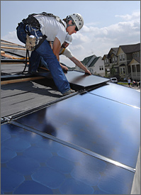 Photo of two men wearing hard hats and safety harnesses installing large solar panels on a roof.  Houses of various sizes are visible in the background.