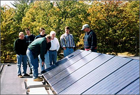 Photo of a high-school faculty member pointing out features of a solar electric system installed on the roof of a New Hampshire high school.  Students and other faculty members stand nearby. Trees are visible in the background.