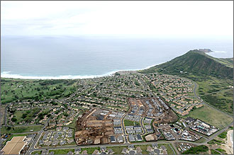 Photo of the Mololani neighborhood in Hawaii. The aerial photo shows a distant view of the neighborhood, which is surrounded by residential and commercial buildings and lush greenery. The Pacific Ocean encompasses the upper half of the photo. 