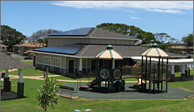 Photo of solar panels on roof of the Halsey Terrace Community Center, which is surrounded by green  grass and a kid's playground.  Clouds and trees can be seen in the background.