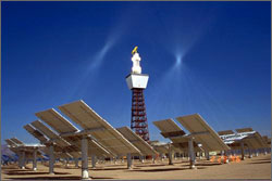 Photo of the central tower of the Solar Two power plant. The upper section containing the receiver is white against a blue sky. In the foreground are numerous large flat mirrors or heliostats that track the sun and focus the light onto the receiver.