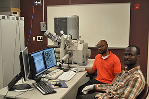 Photo of two males seated in front of a computer with a large machine sitting on a table in the background.