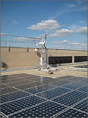 Photo of solar photovoltaic panels lined in four rows on a flat roof. The ledge and railing are in the background.