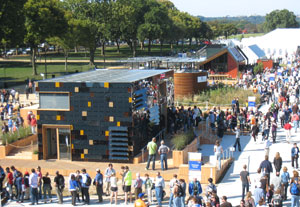 Photo of hundreds of people surrounding a row of small demonstration homes. The exterior of the square-shaped home in the forefront is made of reflective black material.