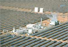 Photo of numerous parallel rows of parabolic trough collectors tracking the sun. The cooling towers and other generator equipment are in the midst of the troughs, and two water tanks are in the background.