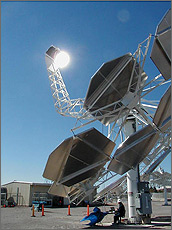 Photo of several flat, octagonal panels arranged together to form a dish-shaped structure. The receiver is supported above the panels by an arm-like structure and glows as it is heated by concentrated sunlight. A technician sits below the dish, in the shade of the panels.