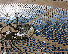 Aerial photo of a power tower system, showing numerous large, reflective mirrors in concentric circular rows. Tracking the sun, each mirror reflects onto the top of the tower at the center of the circle of mirrors. The receiver at the top of the tower is glowing.