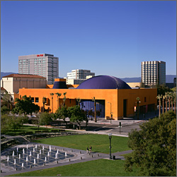 Photo of the blue dome on the roof of the Tech Museum of Innovation in San Jose.  Visible in the foreground are a chessboard-like fountain and grass and in the background are tall buildings framed by blue sky.