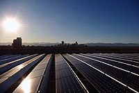 Photo of the sun shining on rows of rooftop solar panels with a skyline and mountains in the background.