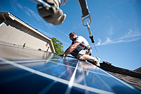 Photo of a man in a harness installing a large blue photovoltaic panel on a rooftop.