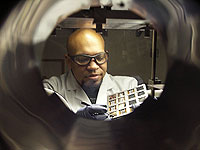 Photo taken through a metal circular vacuum chamber of a lab technician in safety gear manipulating a tray of plastic solar cells.