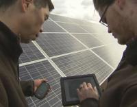 Photo of two men looking at a smartphone and a tablet computer in front of an array of solar panels.