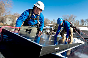 Photo of two males with safety gear mounting a rectagular-shaped solar panel on a roof.