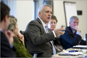 Photo of a man speaking while seated at a table with men and women surrounding him.