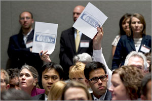 Photo of a room full of people seated in rows with a few standing in the background. Two hands in the crowd are holding up sheets of paper with 'Like'.