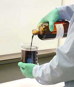 Photo of gloved hands pouring liquid from a glass bottle to glass beaker.