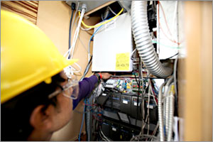 Photo of a man, wearing a yellow hard hat, looking at a wall with panels and wires mounted to it.