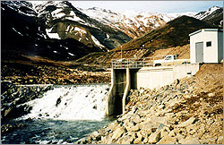 Photograph of rocky, mountainous terrain showing water flowing down a small section of river rapids. To the right of the rapids is a small concrete structure through which part of the river has been diverted. Above the concrete structure is a utility building.