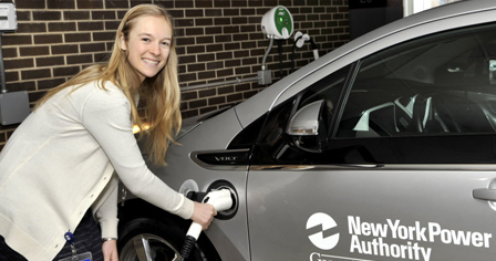 Woman holding the charging unit has she is charging a grey vehicle. Vehicle has New York Power Authority on the driver side door.