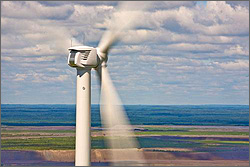Photo of the top of a large wind turbine, its blades spinning, ocean and sky in the background.