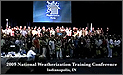 Photograph of the attendees of the National Weatherization Training Conference, lined up in four long rows on a stage. Beneath them, the text reads, '2009 National Weatherization Training Conference, Indianapolis, TN.' They stand in front of a large blue screen with the Weatherization Program's logo on it-a house surrounded by the wind, rain, sun, and snow, above the words 'Weatherization Works.'